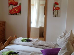 Zimmer mit zwei einzelnen Holz-Betten aus Holz, im Hintergrund eine geöffnete Tür und je ein Bild an der Wand daneben