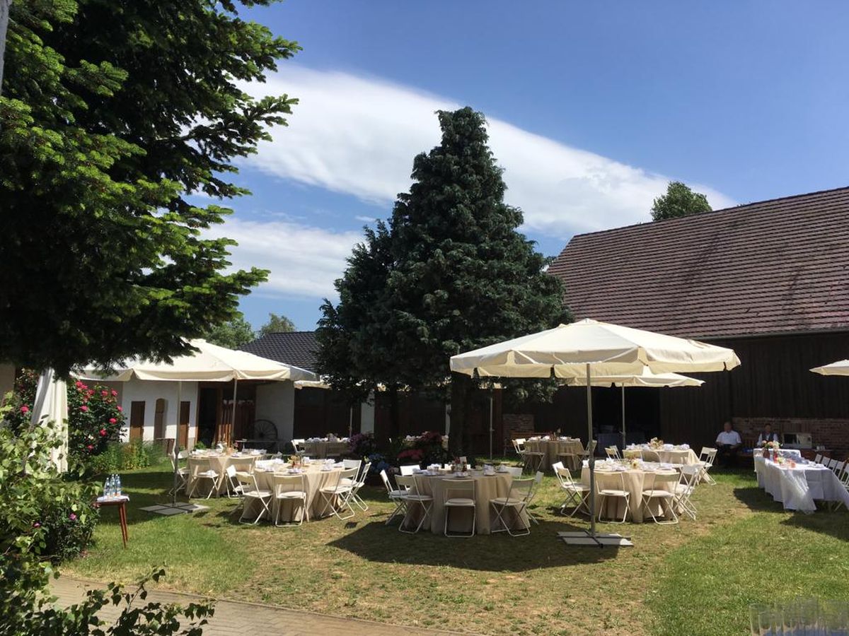 Outdoor: Blick aus dem Garten Richtung Scheune, im Vordergrund Tische, Stühle, Sonnenschirme; im Hintergrund Baum und Nebengebäude